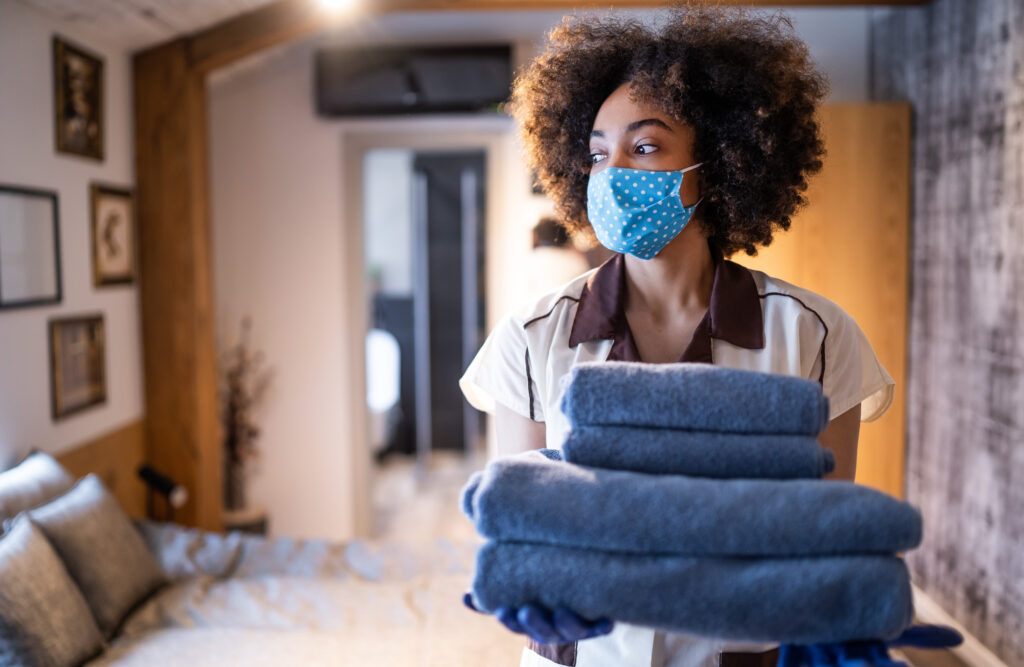 Maid working at a hotel wearing a protective face mask, and holding towels while prepares the hotel room for guests after reopening, during COVID-19