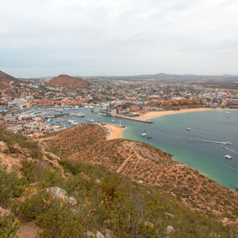 Aerial view of Cabo San Lucas beaches, marina, and buildings surrounded by hills.