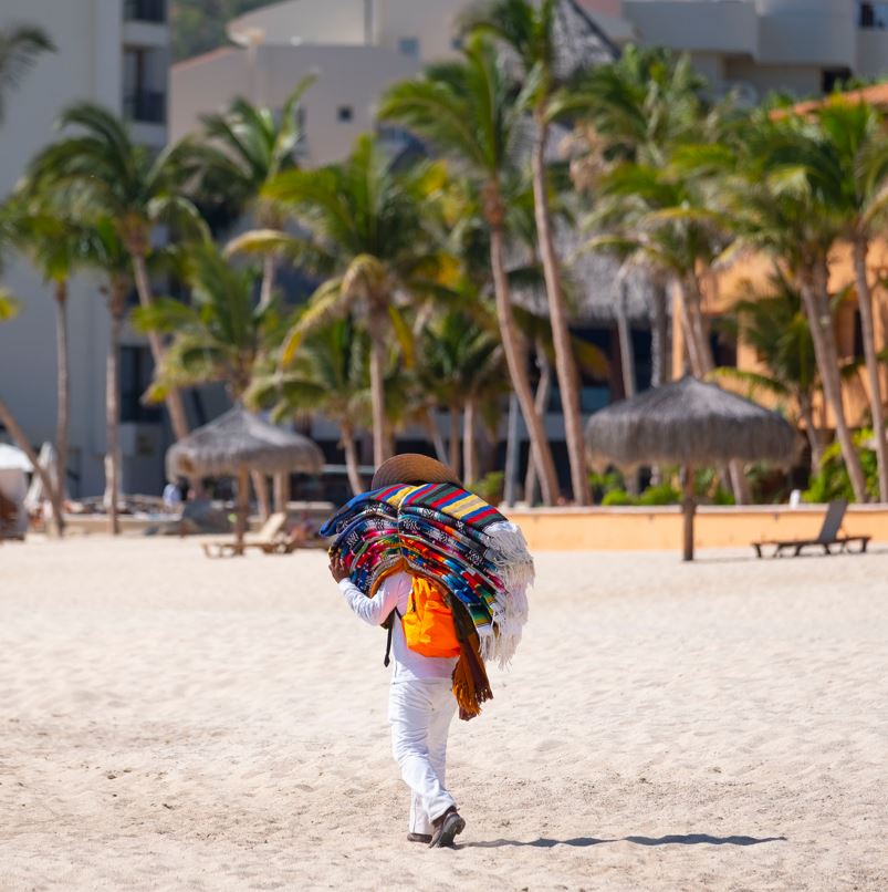 Beach vendor walking on sand with pile of Mexican blankets