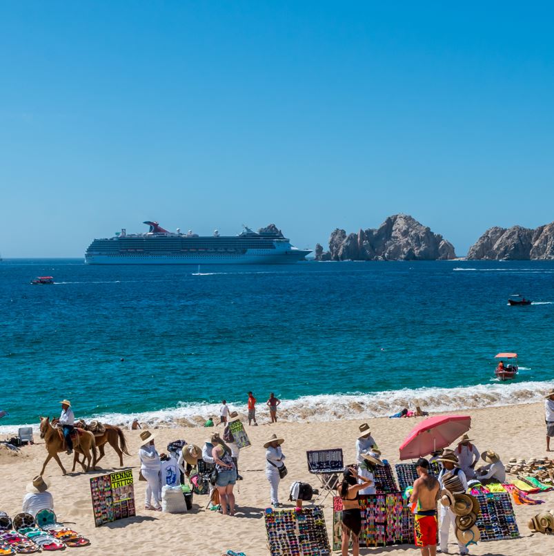 People on a beach in Cabo with a cruise ship on the water in front of the famous arch.