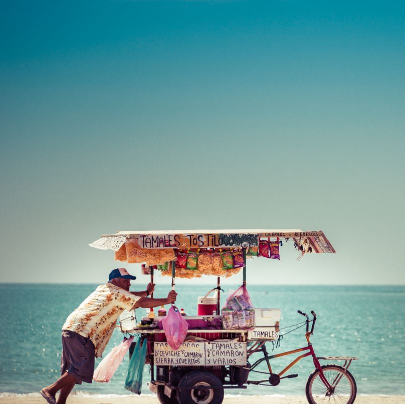 Beach vendor with food and drinks pushing a cart with a bike