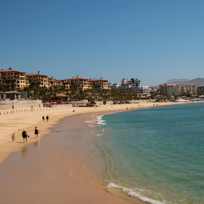 Sandy Cabo Beach with tourist peacefully walking along and buildings in the background.