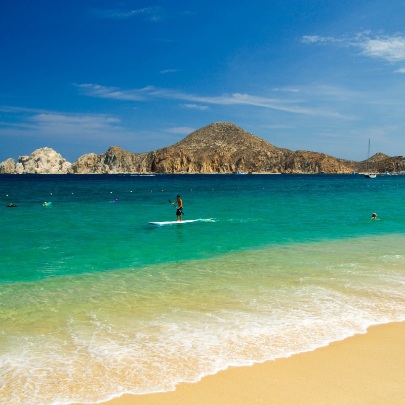 Cabo Beach with a view of Land's End and a person paddleboarding on the turquoise blue water.