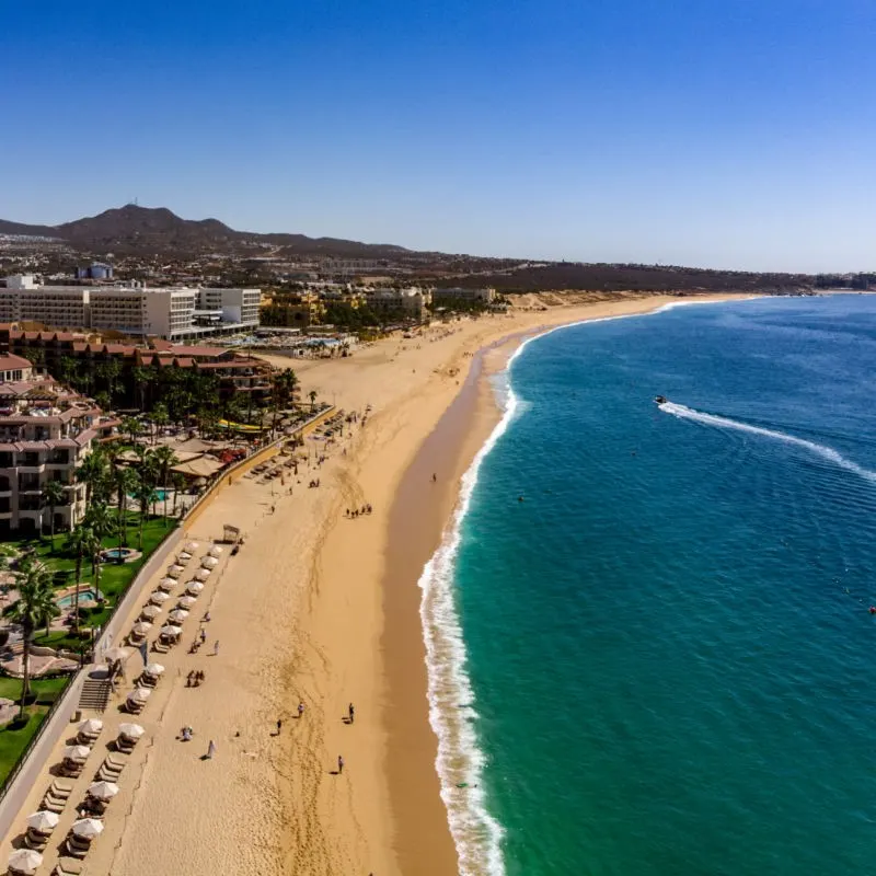 A long stretch of sand on Medano Beach in Cabo San Lucas with hotels in the background.