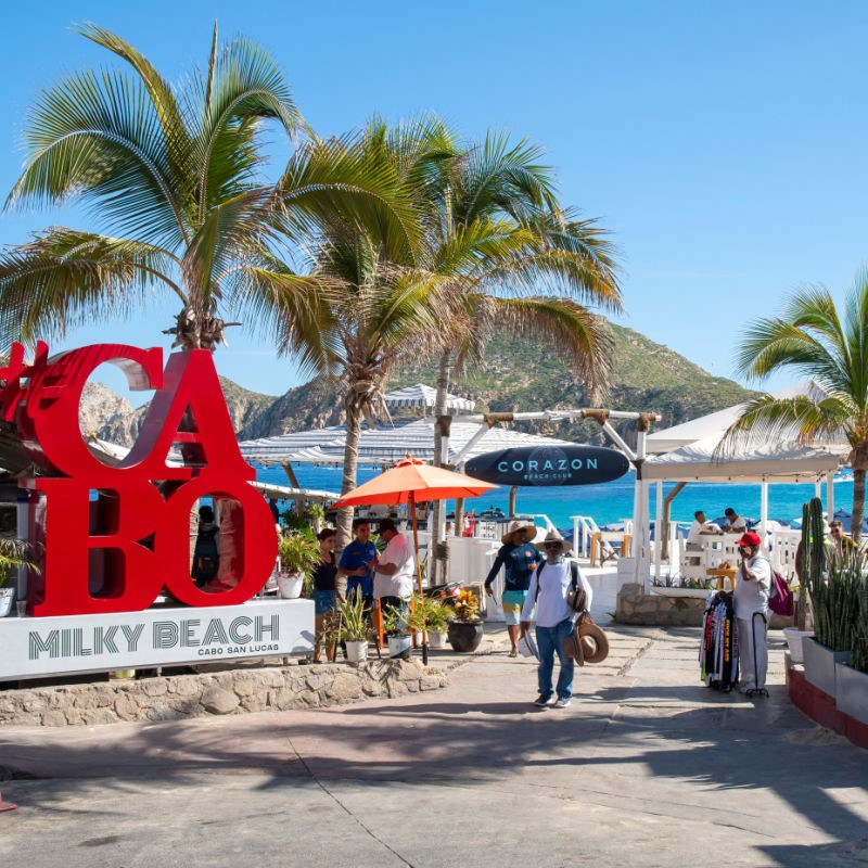Cabo Beach with red sign and people walking towards the beach along a walkway surrounded by palm trees.