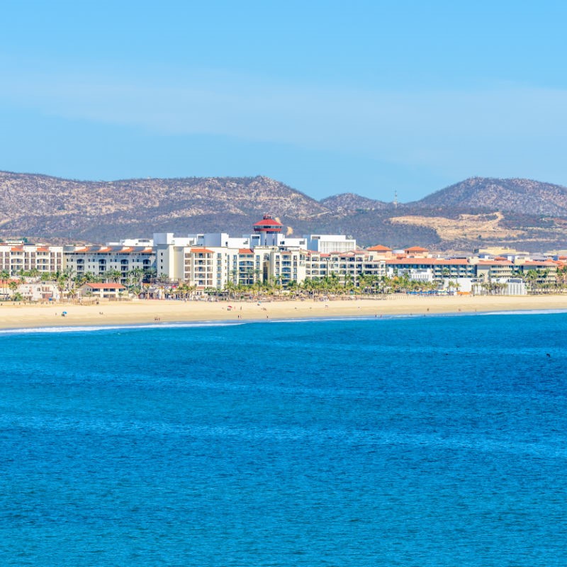 View of San Jose del Cabo from the sea with a sandy beach, resorts, and hills in the background on a sunny day.