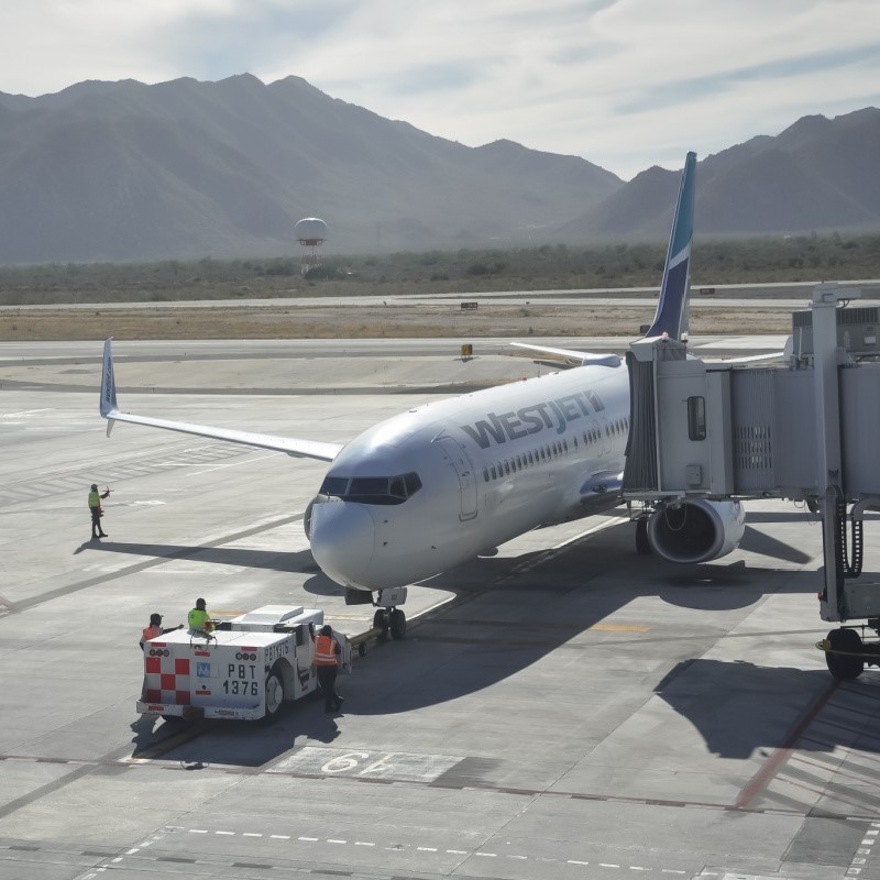 Westjet Plane at Los Cabos Airport on the tarmac with the bridge attached to the plane and workers on the ground with a view of hills in the background.