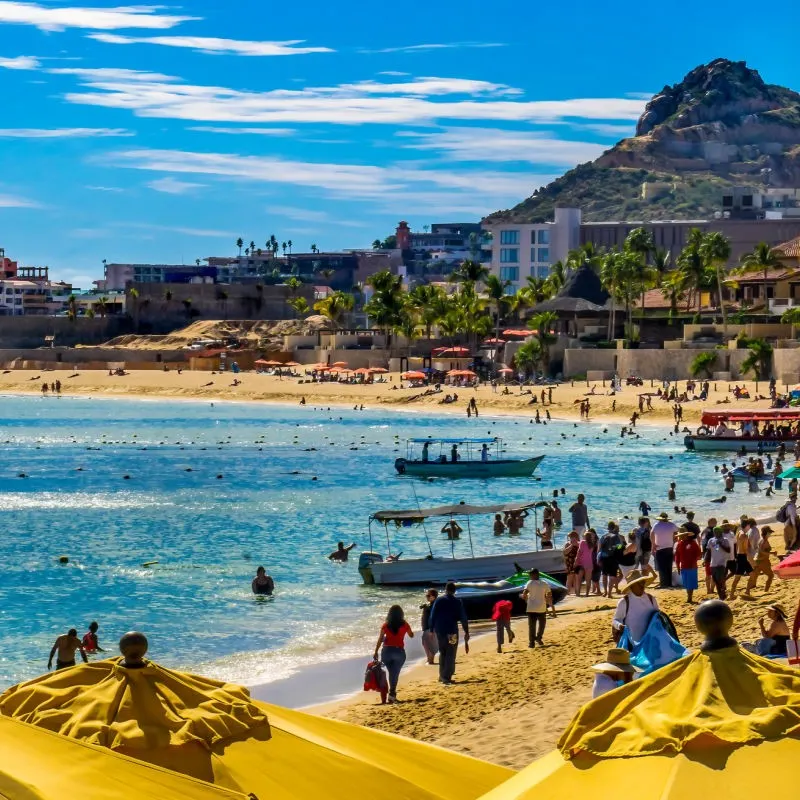 Tourists on the Beach in Los Cabos with boats on the water and sandy beaches and hills in the background.