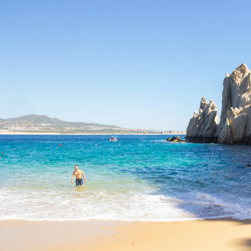 Lover's Beach in Cabo San Lucas with a man walking towards the sandy beach and a backdrop of hills in the distance.