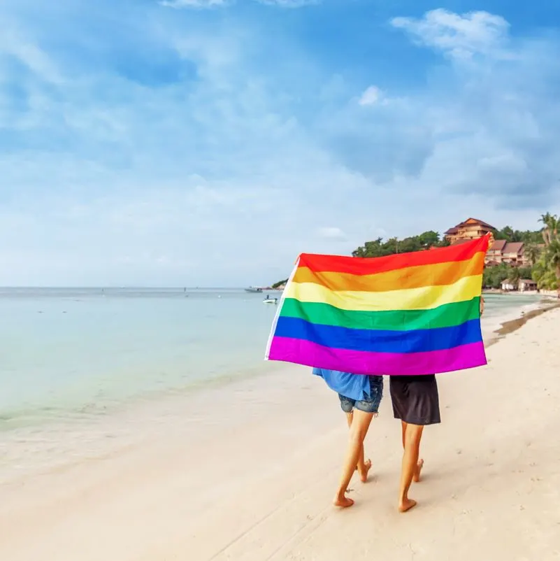 An LGBTQ+ couple walking on a beach with a rainbow flag