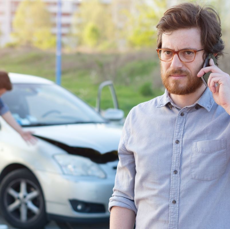 A man using a cell phone with a car in the background thats been in a minor collision