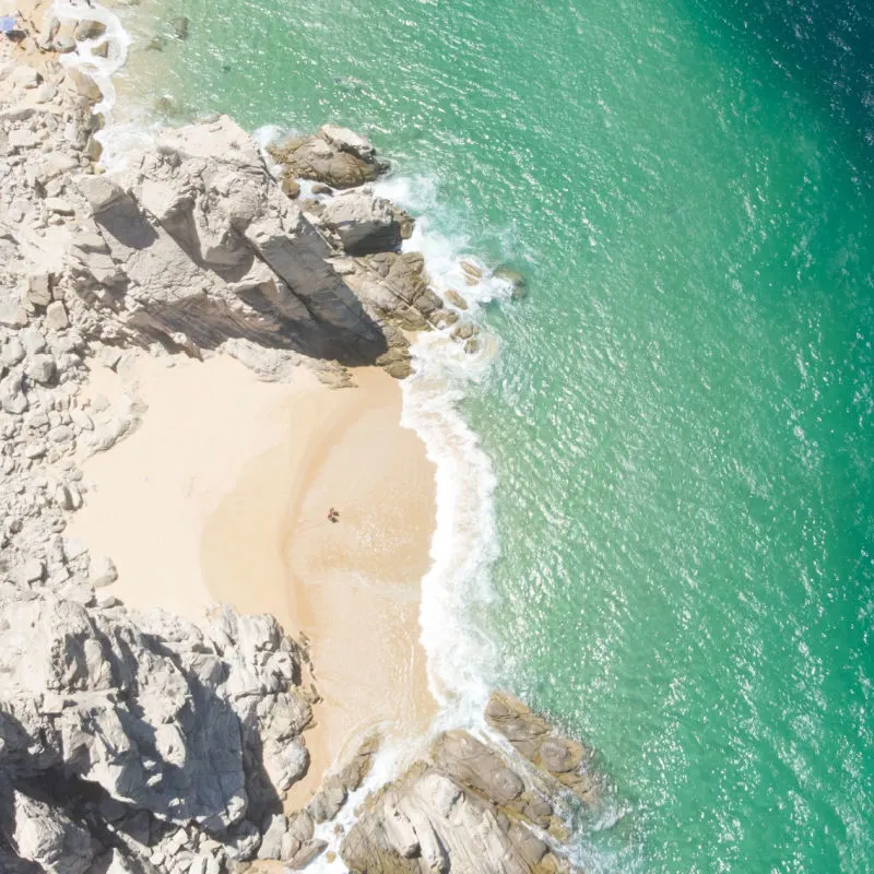 Aerial Image of couple standing on Playa de los Almantes in Cabo San Lucas, Baja California, Mexico. Impressive rocky cliffs and crystal clear turquoise green water with white sand and waves.