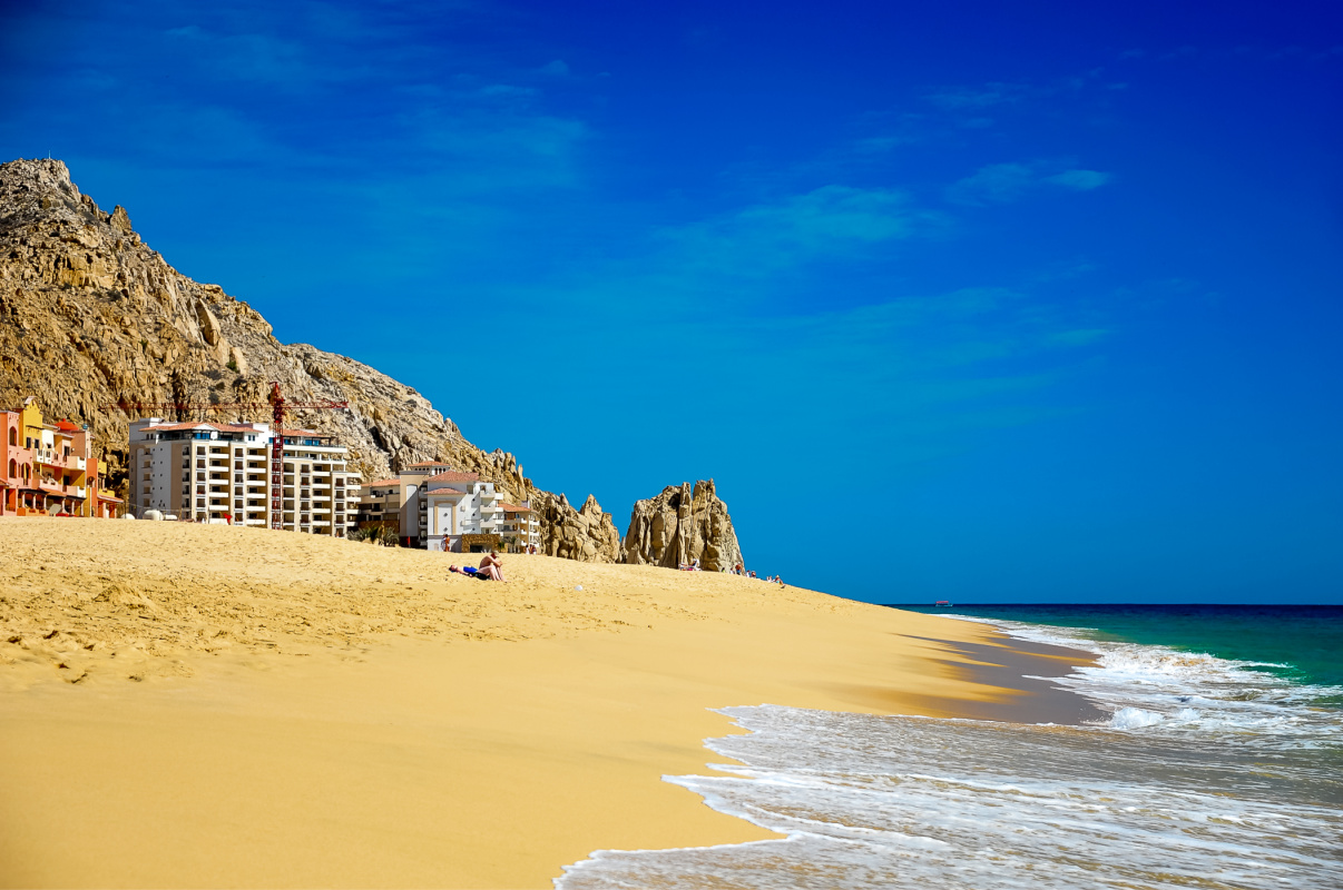 Idyllic beach scene with rocky mountain backdrop, turquoise sea and deep blue sky. Bathers relaxing on Pedregal beach, Cabo San Lucas, Mexico