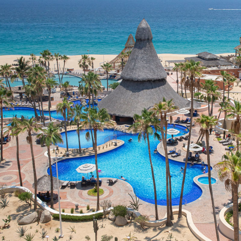 a beach resort in los cabos with multiple pools and a palm lined palapa