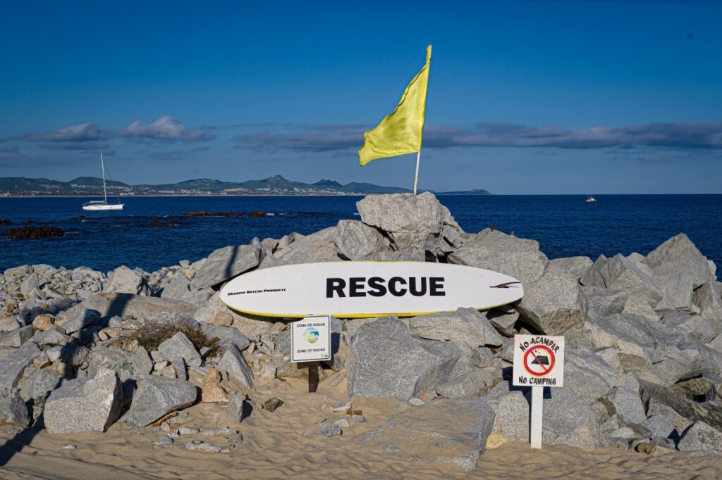 Photo of lifeguard rescue surfboard with yellow warning flag and no camping sign at Palmilla Beach