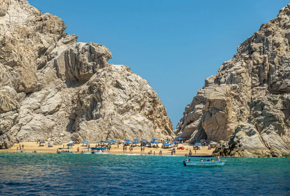 Narrow sandy patch between rocky cliffs leading to other side, the Playa de los Amantes. Blue umbrellas and sunbathers under blue sky. Glass bottom boat