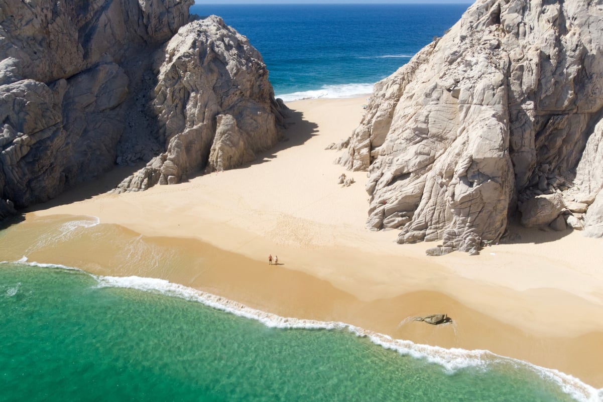 Aerial Image of couple standing on Playa de los Almantes in Cabo San Lucas, Baja California, Mexico. Impressive rocky cliffs and crystal clear turquoise green water with white sand at lover beach.